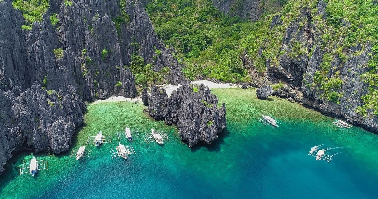 An aerial view of outrigger boats floating on turquoise water in a secluded cove with jagged limestone cliffs and a white sand beach.