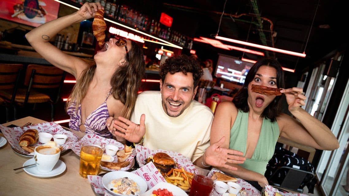 Un voyage WeRoad en groupe de trois personnes profitant d'un brunch amusant, faisant des grimaces avec un croissant et du bacon dans un restaurant.