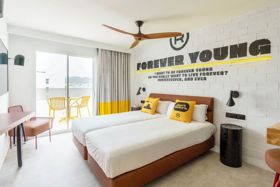 Una habitación de hotel moderna con dos camas individuales, almohadas decorativas amarillas y un mural "Forever Young" en una pared de ladrillo blanco.