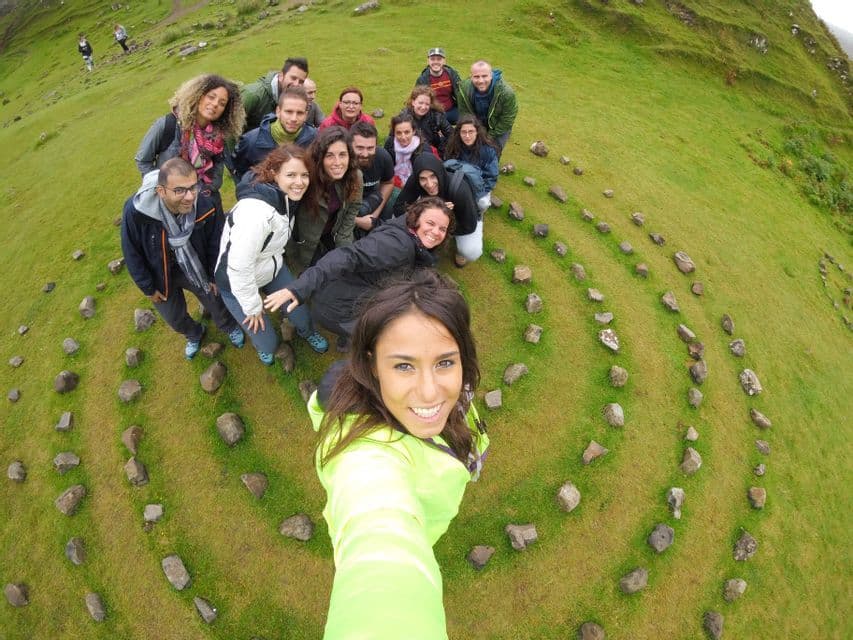 Un gruppo WeRoad in viaggio si scatta un selfie all'interno di una formazione a spirale in pietra su una vivace collina verde.