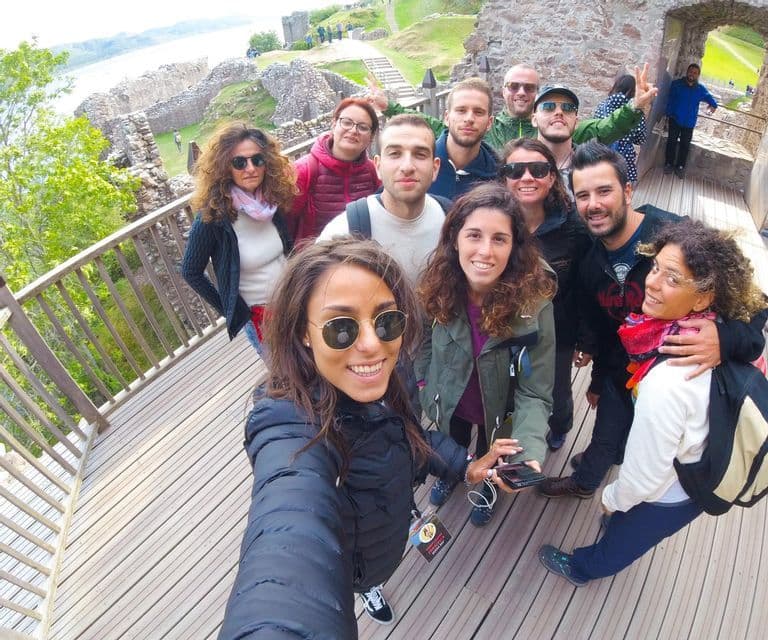 Un groupe WeRoad souriant prend un selfie sur une passerelle en bois, avec des ruines de château en pierre en arrière-plan.