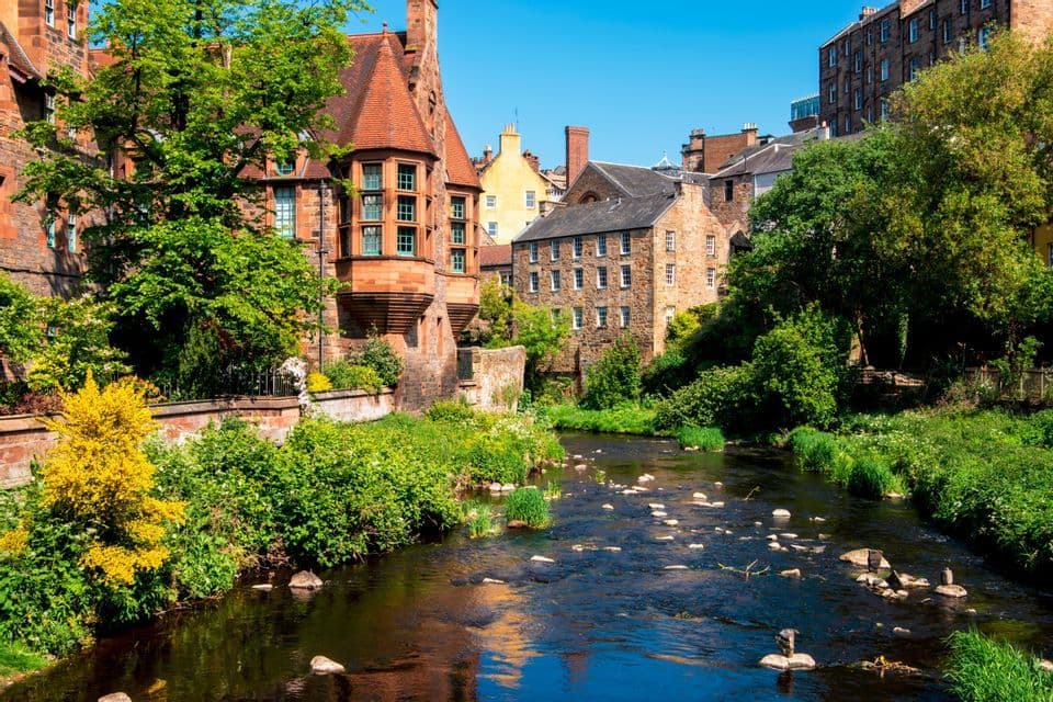 Une rivière traverse un village historique, ses rives bordées de bâtiments en pierre traditionnels et d'arbres verts luxuriants, sous un ciel bleu clair.
