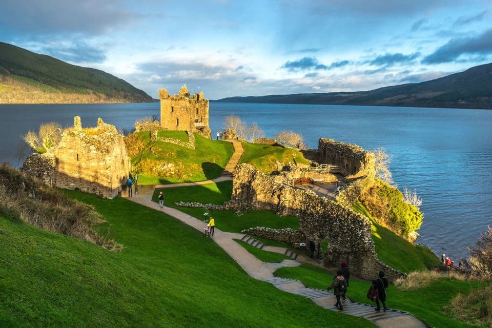 Les ruines d'un château de pierre se dressent sur une colline herbeuse surplombant un grand lac, avec des gens se promenant le long des sentiers.
