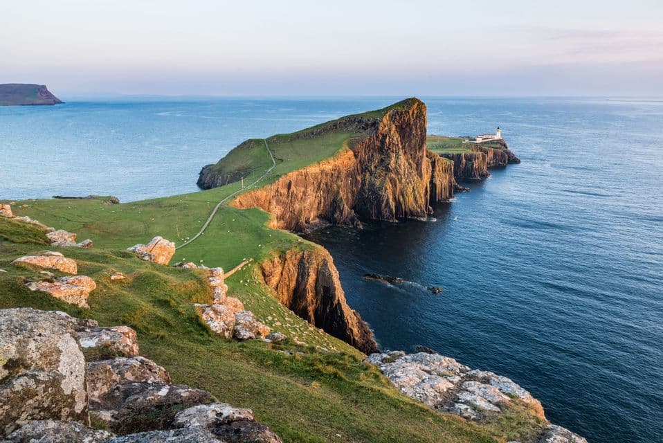 Un faro bianco si erge su una penisola verde a picco che si protende in un mare blu calmo sotto un cielo sereno.