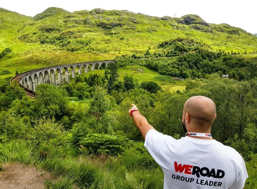 Un accompagnatore WeRoad, visto da dietro, indica un grande viadotto in pietra che si snoda attraverso una lussureggiante valle verde e colline.