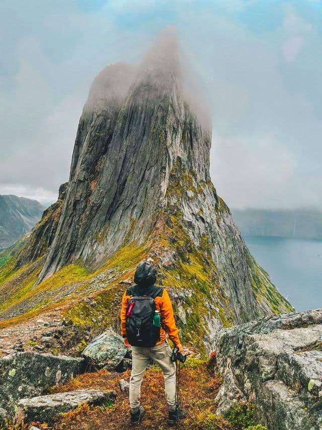 Un randonneur avec un sac à dos et une veste orange se tient sur une crête moussue, regardant vers un pic rocheux abrupt partiellement couvert par les nuages.