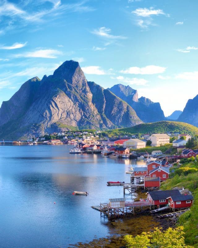A seaside village with red houses on stilts sits on a calm bay at the foot of towering mountains under a blue sky.
