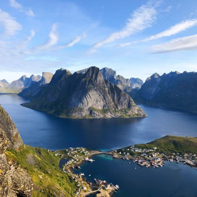 Vue aérienne d'un village côtier entouré d'un fjord d'un bleu profond et de montagnes rocheuses escarpées sous un ciel partiellement nuageux.
