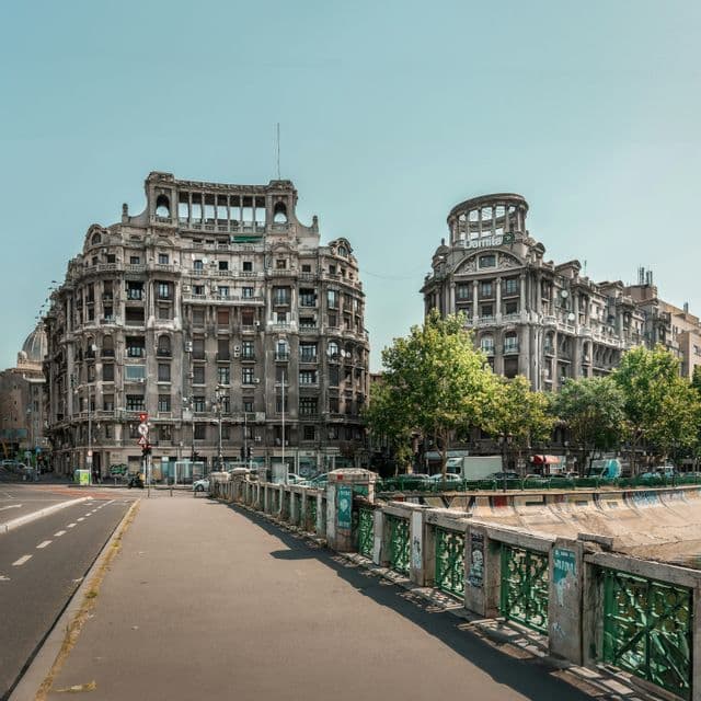 A sidewalk follows a river canal with a green railing, leading towards two large historic buildings under a clear sky.