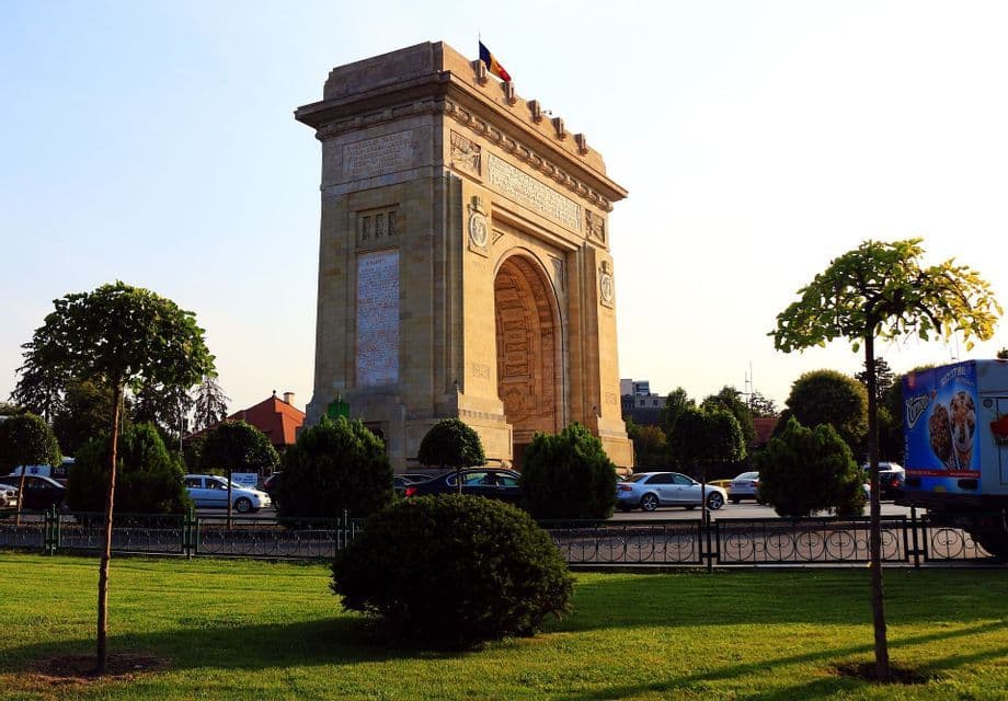 A large stone triumphal arch with a flag on top, viewed from a green park with traffic on a road in the background.