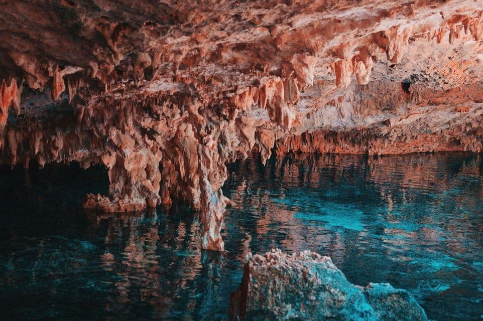 Reddish-orange stalactites hang from the ceiling of a cave above a pool of clear, turquoise water.