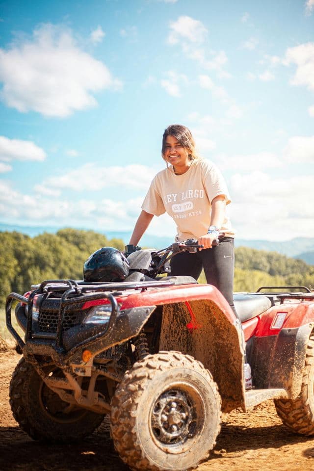 A smiling woman holds the handlebars of a red, muddy ATV on a dirt track, with green hills and a blue sky in the background.