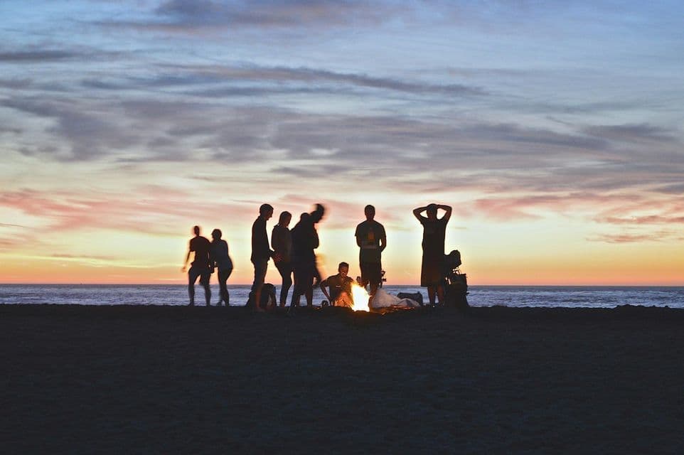 Silhouettes of a WeRoad group trip gathered around a bonfire on a beach at sunset, with the ocean in the background.