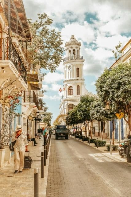 A narrow, tree-lined city street with historic buildings leads towards a white clock tower under a partly cloudy sky.