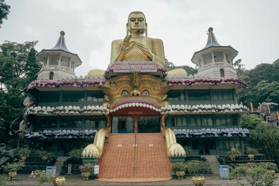 Una grande statua dorata di Buddha si trova in cima a un tempio ornato con un ingresso a forma di bocca aperta di una creatura.