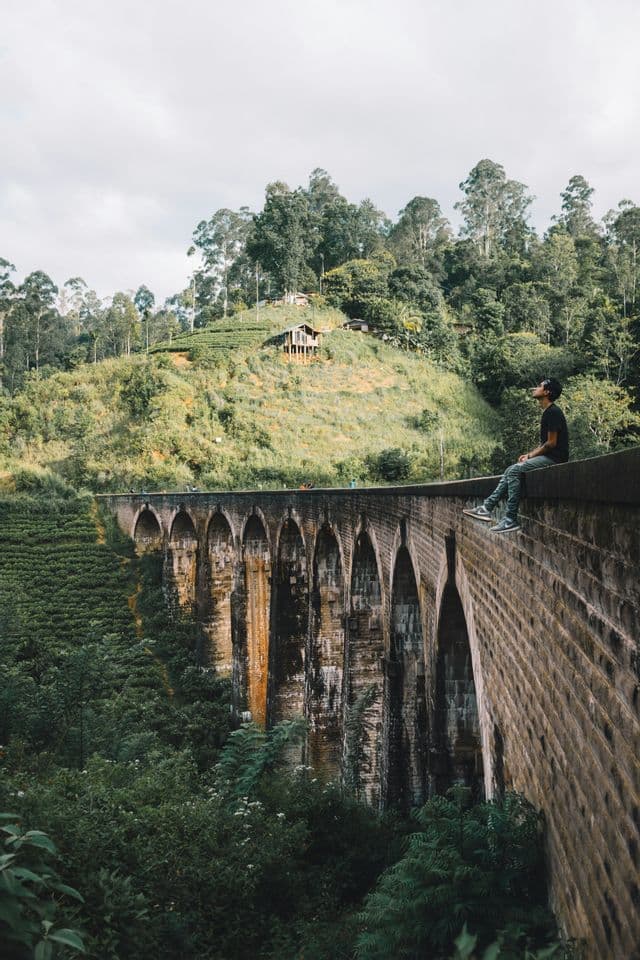 Un uomo in maglietta nera siede sul bordo di un grande ponte ad arco in pietra, guardando una rigogliosa collina verde e boscosa.