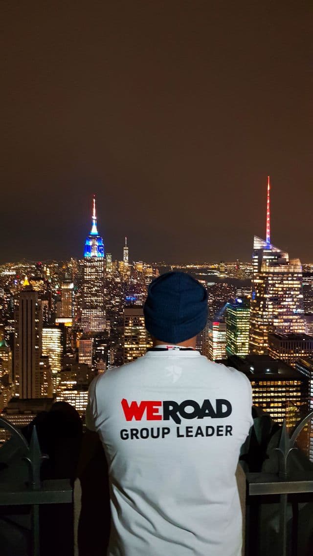 Eine Person von hinten, die ein WeRoad Group Leader T-Shirt und eine blaue Mütze trägt, blickt bei Nacht auf eine beleuchtete Stadtsilhouette.