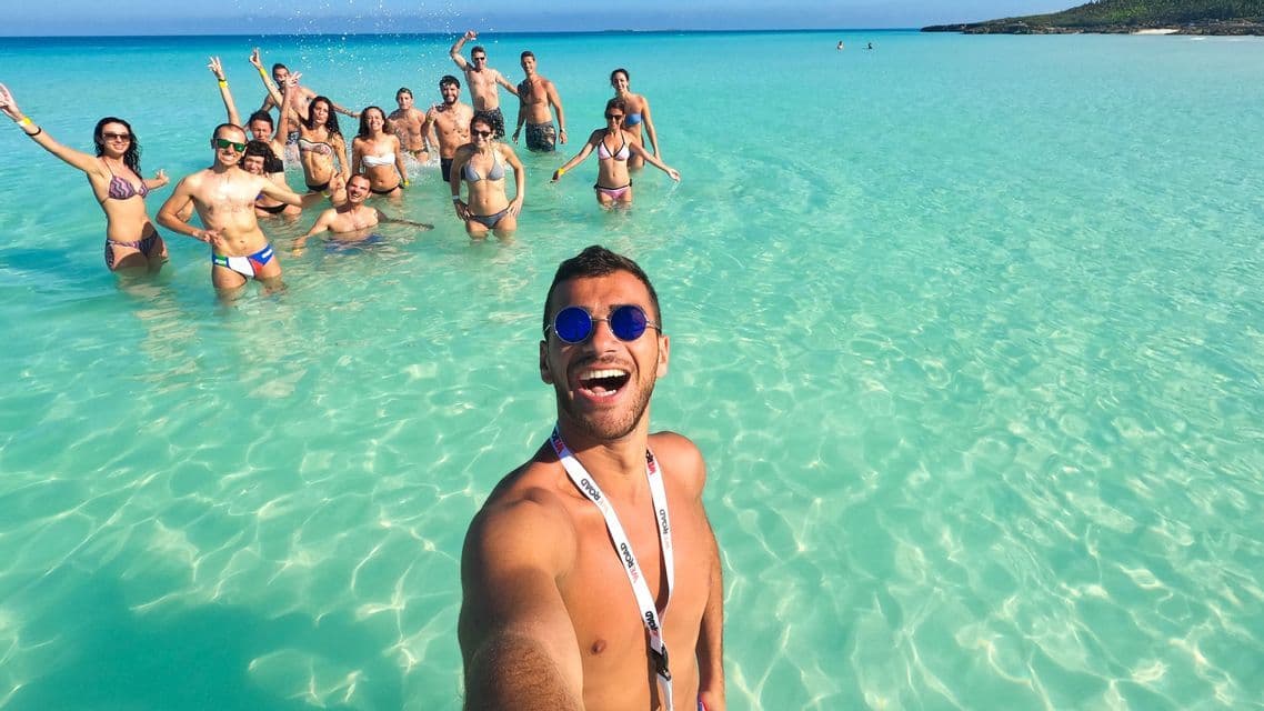 Ein Mann macht ein Selfie mit seiner WeRoad Gruppenreise, alle lächeln und stehen in klarem türkisfarbenem Wasser.