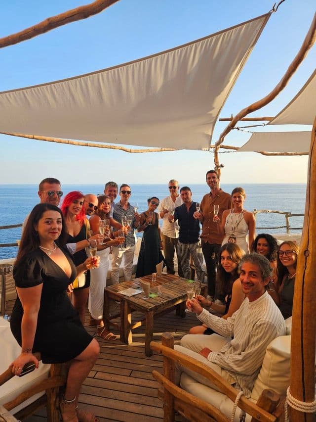 Un voyage de groupe WeRoad avec des personnes trinquant au champagne sur une terrasse en bois surplombant l'océan.