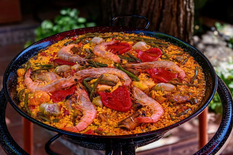 Una padella di paella con riso giallo, gamberi, peperoni rossi e fagiolini, poggiata su un supporto all'aperto.