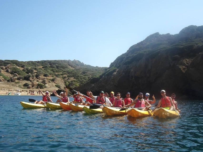 A WeRoad group trip wearing red life vests sits in yellow kayaks, posing on the sea with a rocky coast behind them.