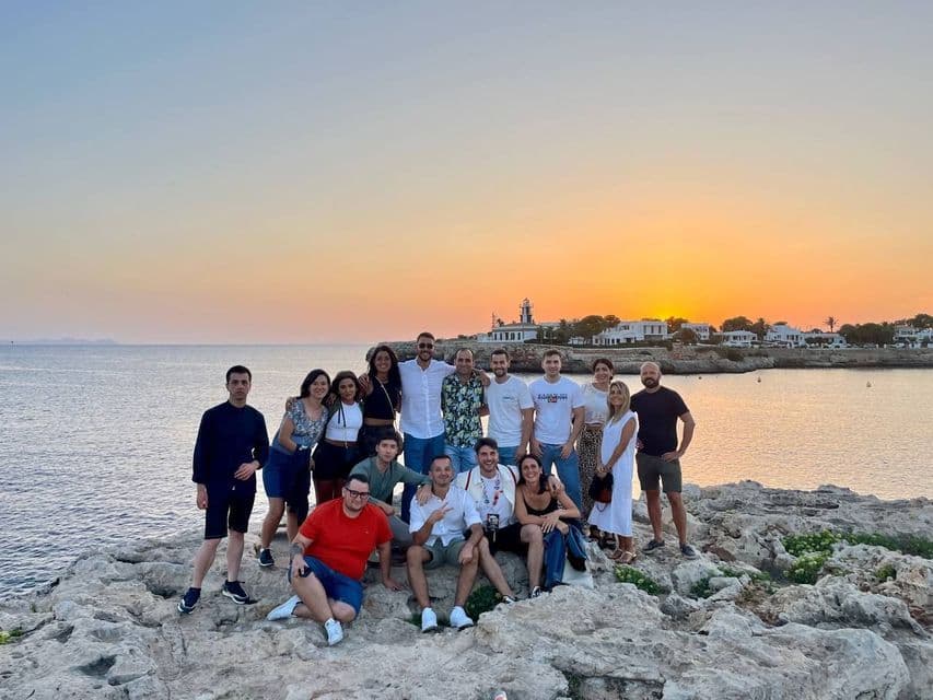 A WeRoad group trip posing for a photo on a rocky shoreline by the sea during a golden sunset, with a lighthouse in the distance.