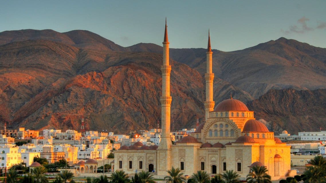 A large mosque with two minarets glows in the golden light of sunset, with a city and rugged mountains in the background.