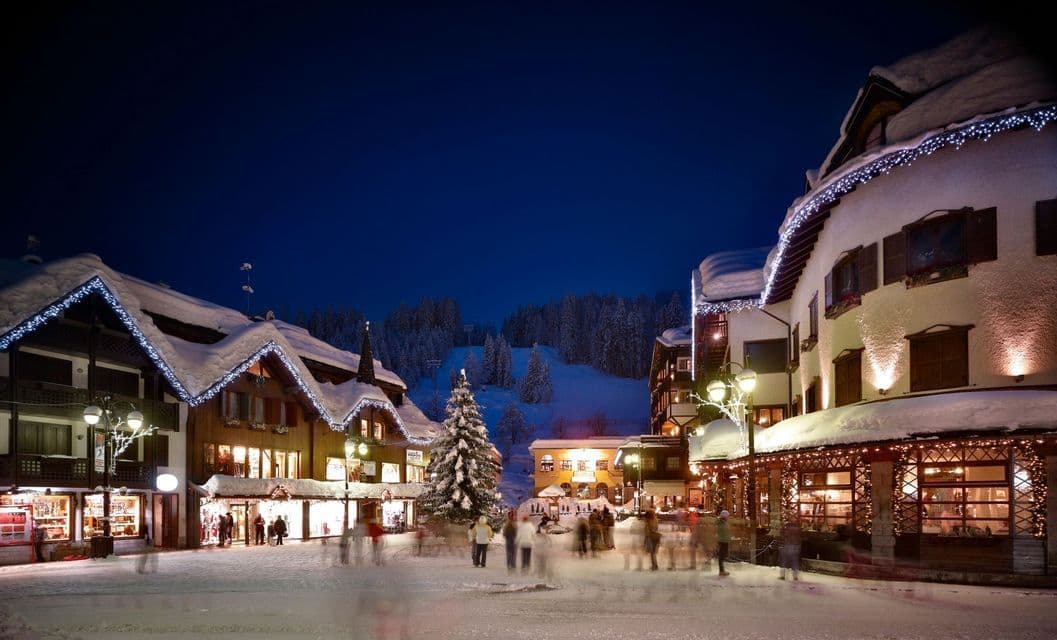 A snowy village square at night, with people walking among chalets and a tree decorated with bright lights under a dark sky.