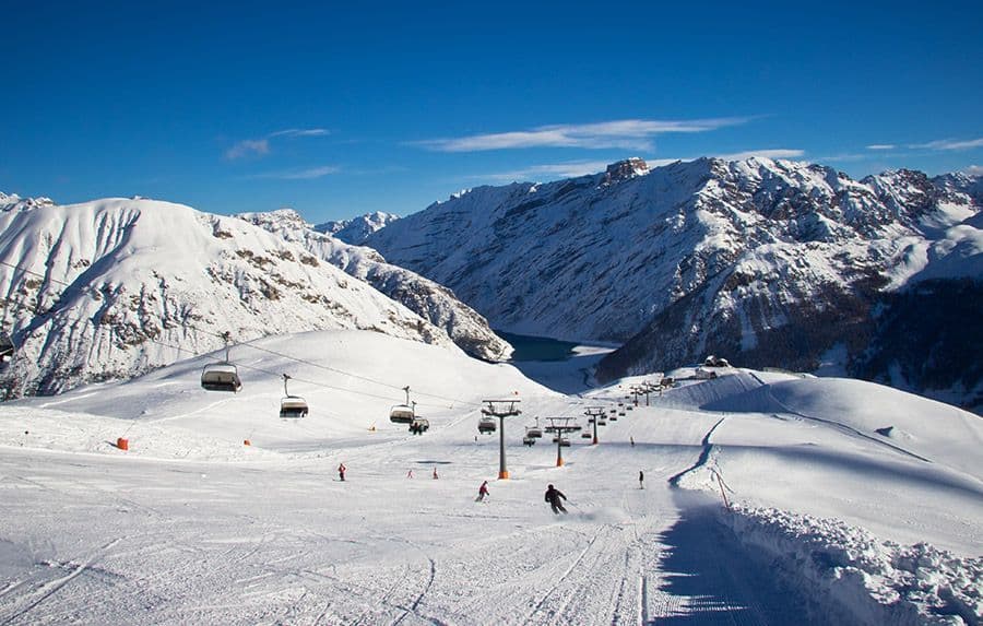 A wide view of a ski slope with people skiing, chairlifts, and snow-covered mountains under a clear blue sky.
