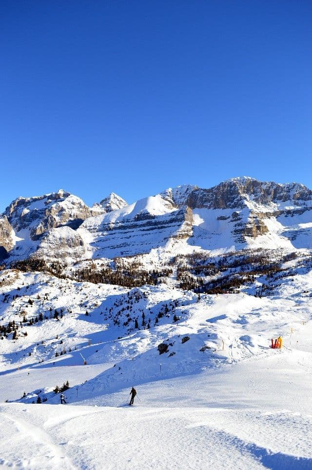 A person skis down a wide, sunny slope against a backdrop of snow-covered mountains under a clear blue sky.