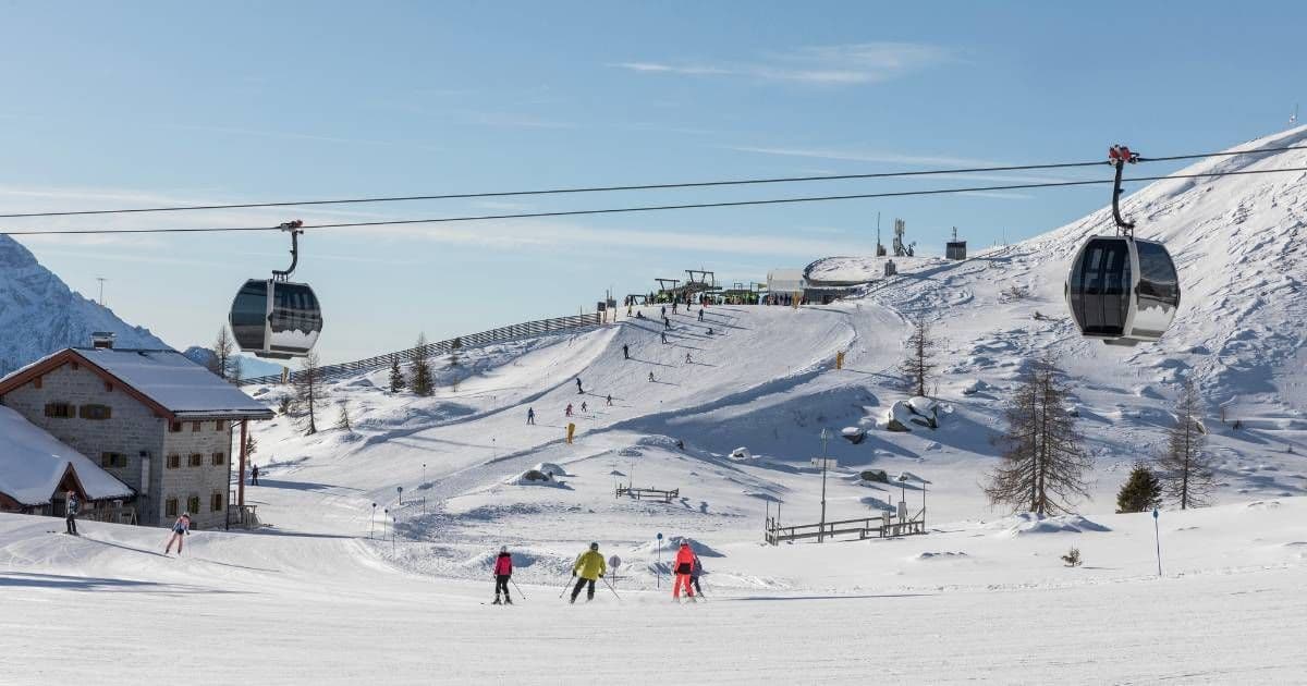 Cable car cabins move over a busy ski slope with people skiing past a stone building under a clear blue sky.