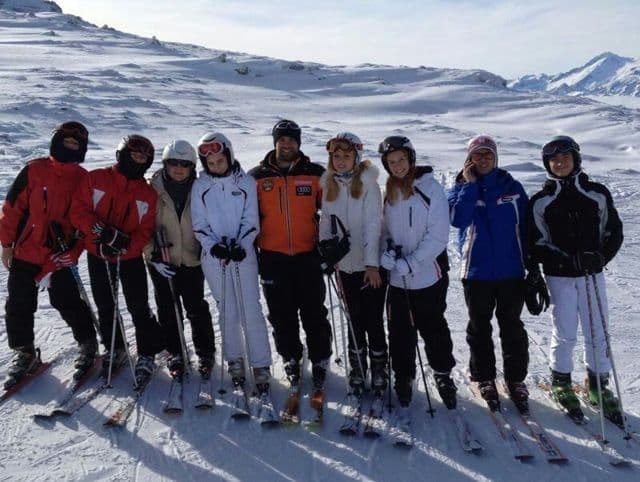 A WeRoad group trip wearing full ski gear poses for a group photo on a snow-covered mountain slope under a clear sky.