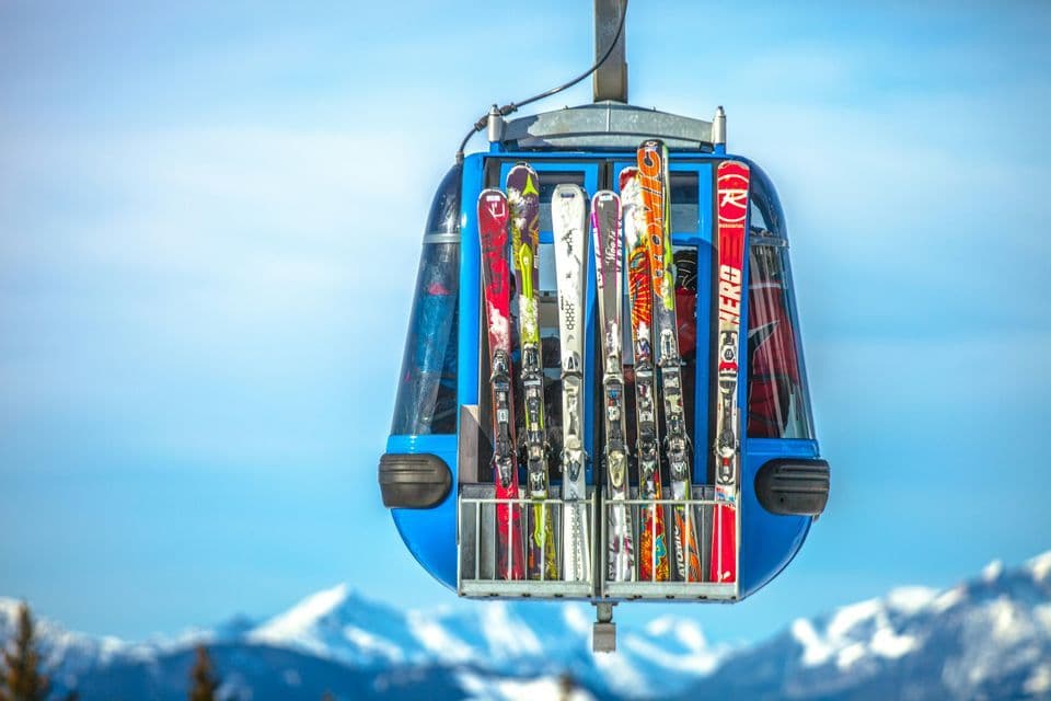 A close-up of skis secured in a rack on the side of a blue cable car gondola, with snowy mountains in the background.