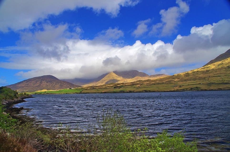 Un lac bleu foncé se trouve au premier plan avec des collines verdoyantes et ensoleillées à l'arrière-plan sous un ciel bleu parsemé de nuages blancs.