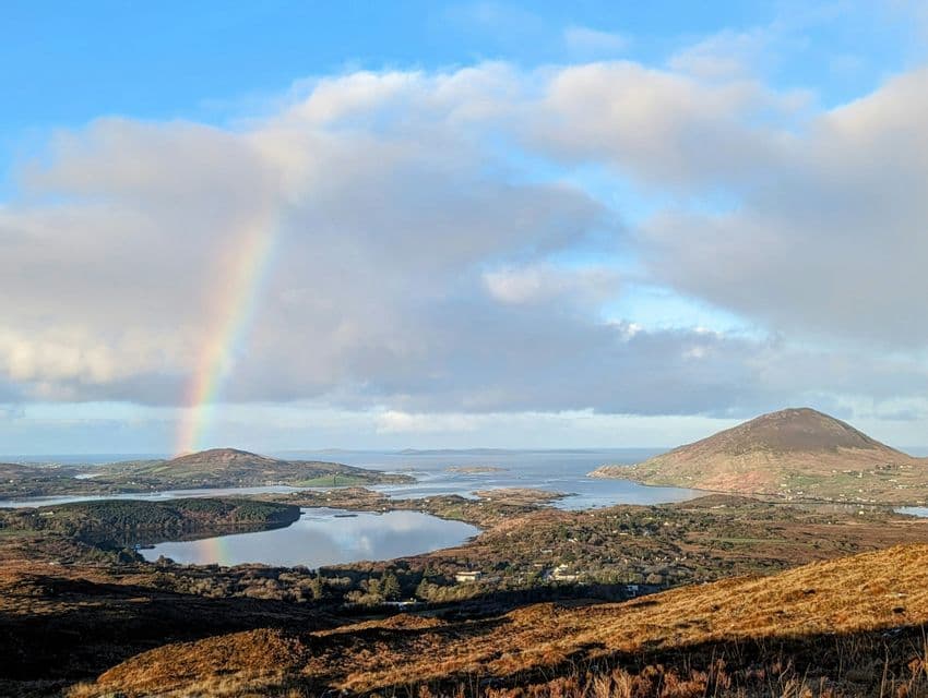 Un arc-en-ciel enjambe un paysage côtier de collines et de lacs, se reflétant dans l'eau sous un ciel partiellement nuageux.