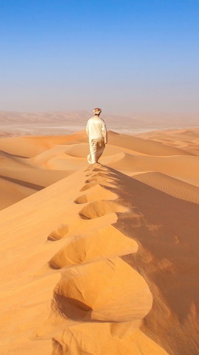 A person in traditional robes walks along the ridge of a large sand dune in the desert, leaving footprints behind.