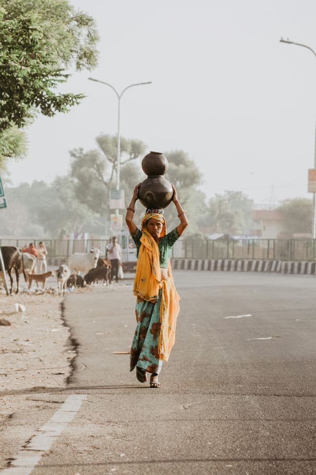 Une femme en sari coloré porte deux pots superposés sur sa tête en marchant sur une route pavée.