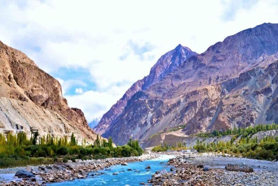 Une rivière bleu vif coule à travers une vallée rocheuse, flanquée d'arbres verts et de montagnes imposantes et escarpées sous un ciel nuageux.