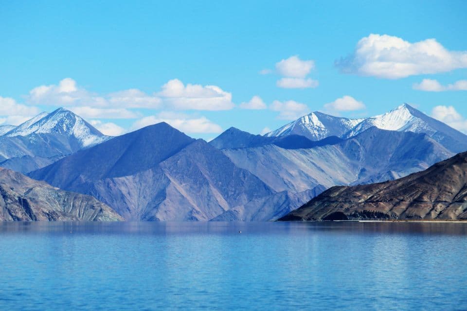 Derrière un lac bleu paisible s'élève une chaîne de montagnes rocheuses aux sommets enneigés, sous un ciel parsemé de nuages blancs.