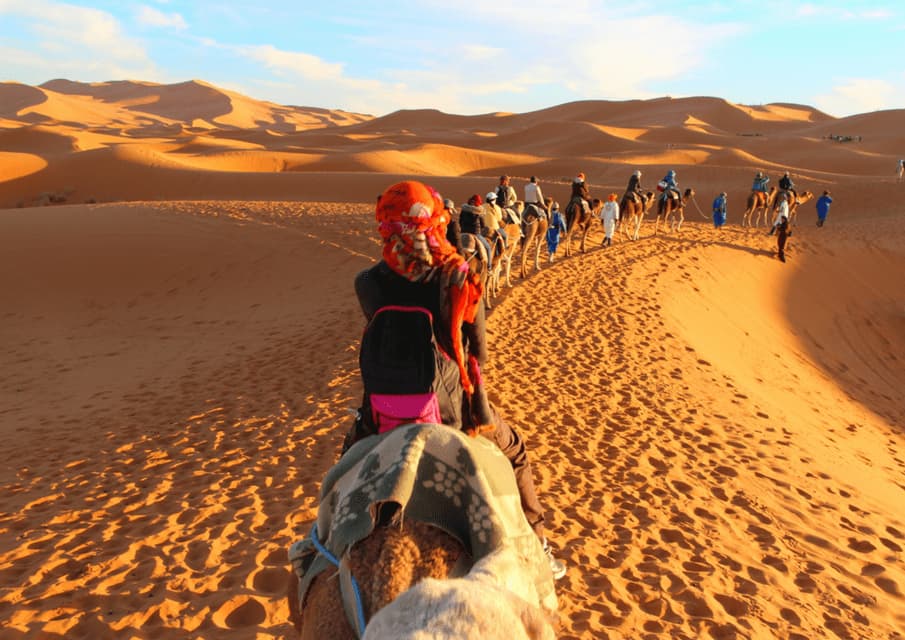 Un viaje en grupo de WeRoad montando camellos en fila india a través de vastas dunas de arena del desierto al atardecer