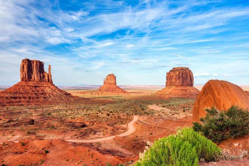 Ampia vista di tre butte di arenaria rossa in un paesaggio desertico sotto un cielo azzurro parzialmente nuvoloso, con una strada sterrata sottostante.