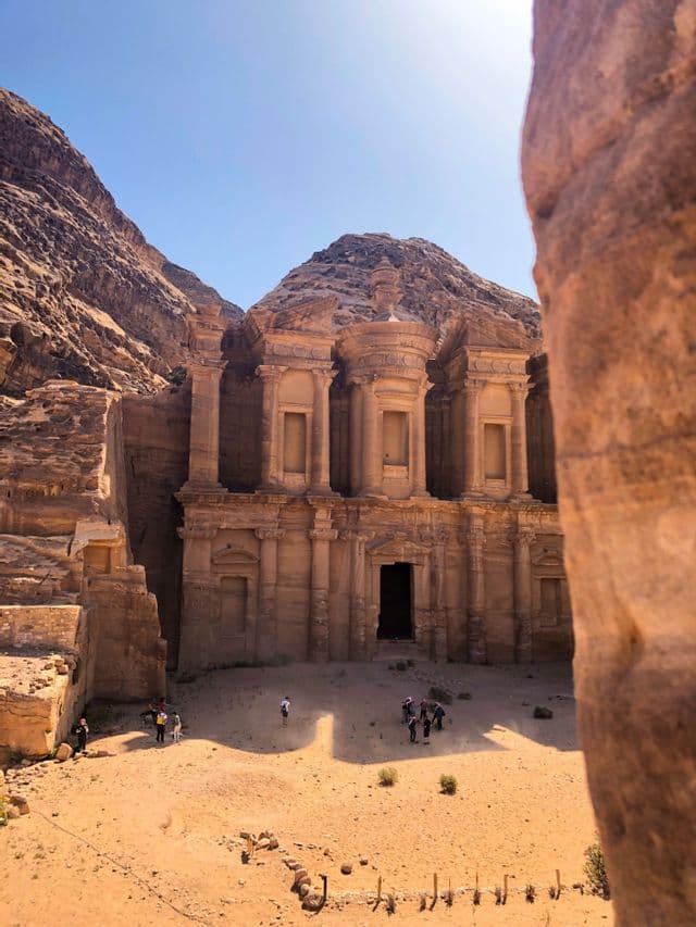 A WeRoad group trip stands in front of an ancient temple carved into a desert rock face under a clear blue sky.