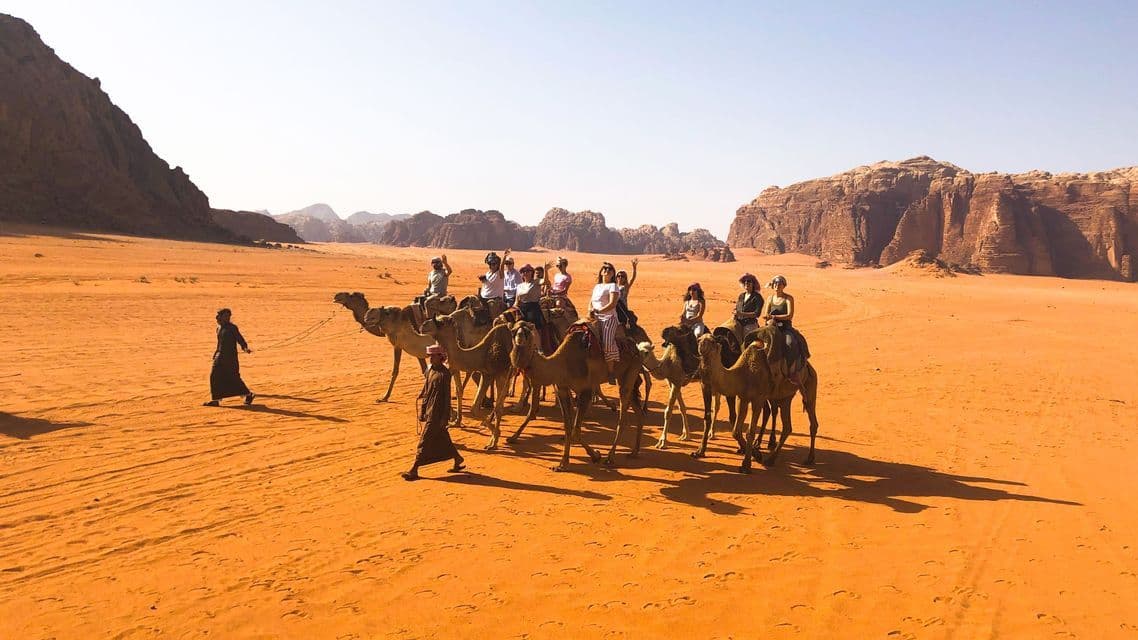 A WeRoad group trip rides on camels through a red sand desert with rocky mountains in the background.