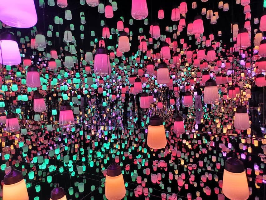 A room filled with hundreds of hanging lanterns glowing in shades of pink and teal, reflected infinitely by mirrored walls.