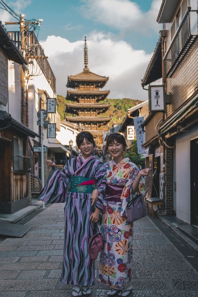 Due donne in kimono colorati sorridono su una strada lastricata con una pagoda tradizionale giapponese visibile sullo sfondo.