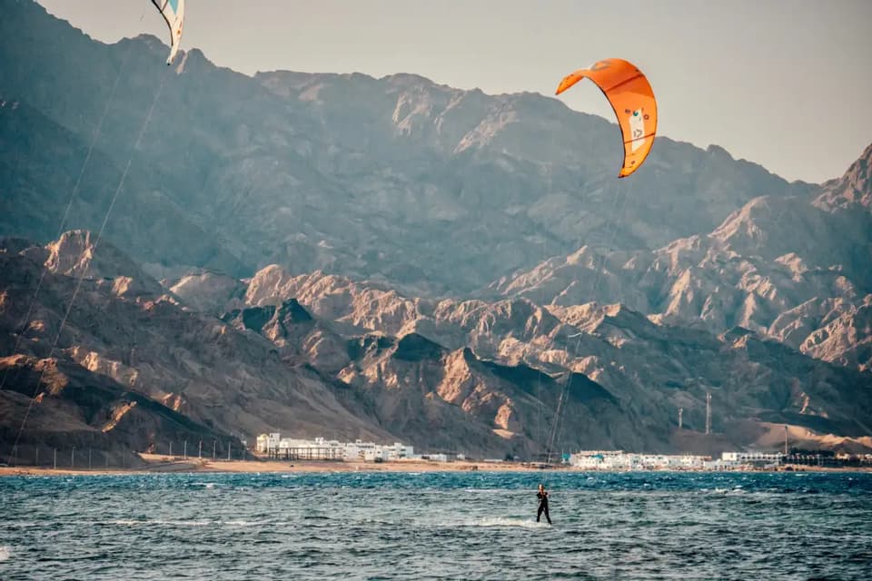 A person kitesurfing on the sea with an orange kite, with a rocky mountain range and coastal buildings in the background.