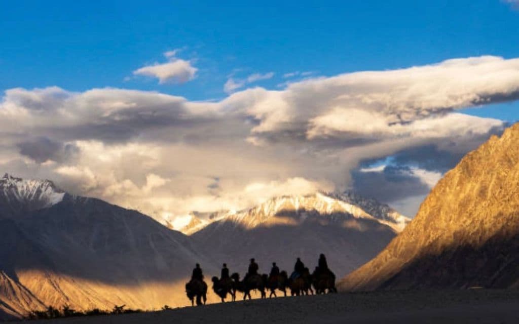 Voyage de groupe WeRoad : silhouettes de chameaux traversant un désert avec montagnes enneigées et ensoleillées.