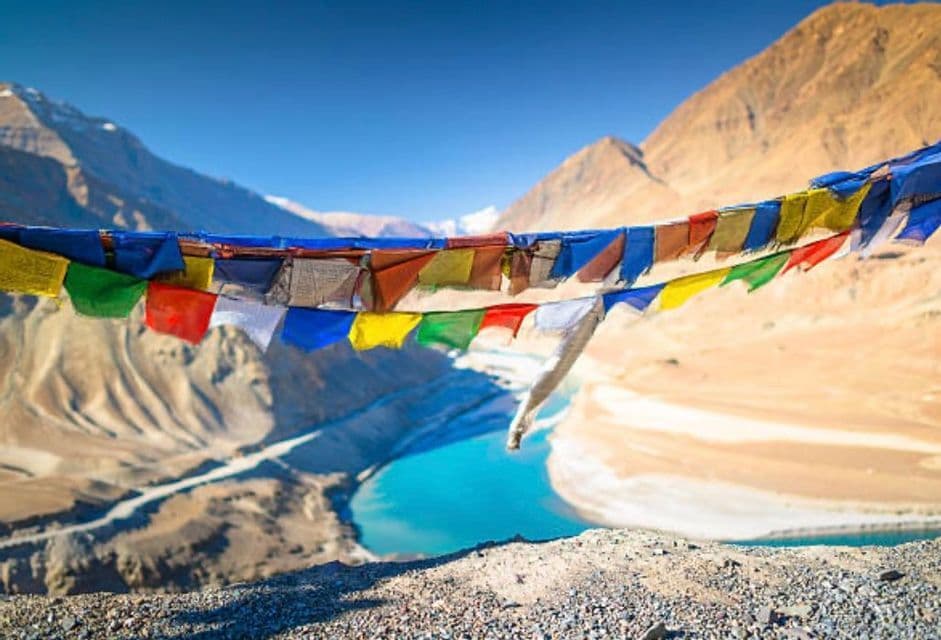 Coloridas banderas de oración cuelgan sobre un río turquesa que serpentea por un valle montañoso árido.