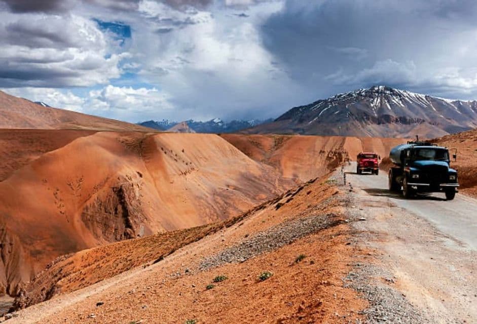 Un'autocisterna blu e un camion rosso percorrono una strada sterrata tortuosa tra montagne rossastre con cime innevate in lontananza.