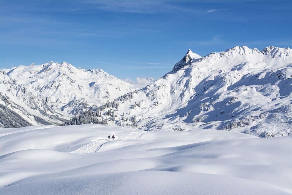 Due escursionisti seguono un sentiero attraverso un vasto paesaggio montano innevato sotto un cielo azzurro brillante.
